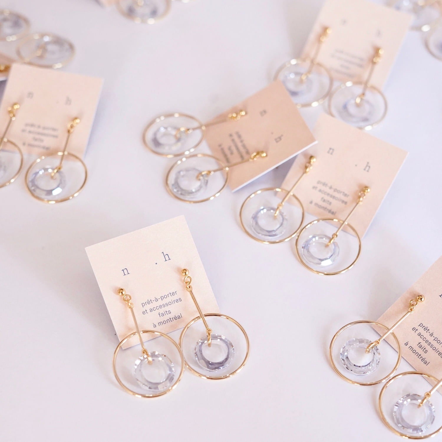 Several pairs of Céleste Earrings by Noémiah, featuring gold-plated hoops with clear Swarovski crystal accents, are showcased on white cards adorned with printed text. The layout on a white surface emphasizes the striking yet sophisticated design of these versatile jewelry pieces.