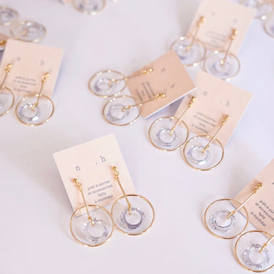 Several pairs of Céleste Earrings by Noémiah, featuring gold-plated hoops with clear Swarovski crystal accents, are showcased on white cards adorned with printed text. The layout on a white surface emphasizes the striking yet sophisticated design of these versatile jewelry pieces.