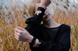 In a field of tall grass, a person with short blonde hair strikes a pose that evokes an autumnal feel reminiscent of the elegance associated with Noémiah's Célia Butterfly Dress. Their eyes are closed, and their right arm is raised to their forehead, while they wear a black silk cotton top adorned with butterfly embroidery on the cuffs.