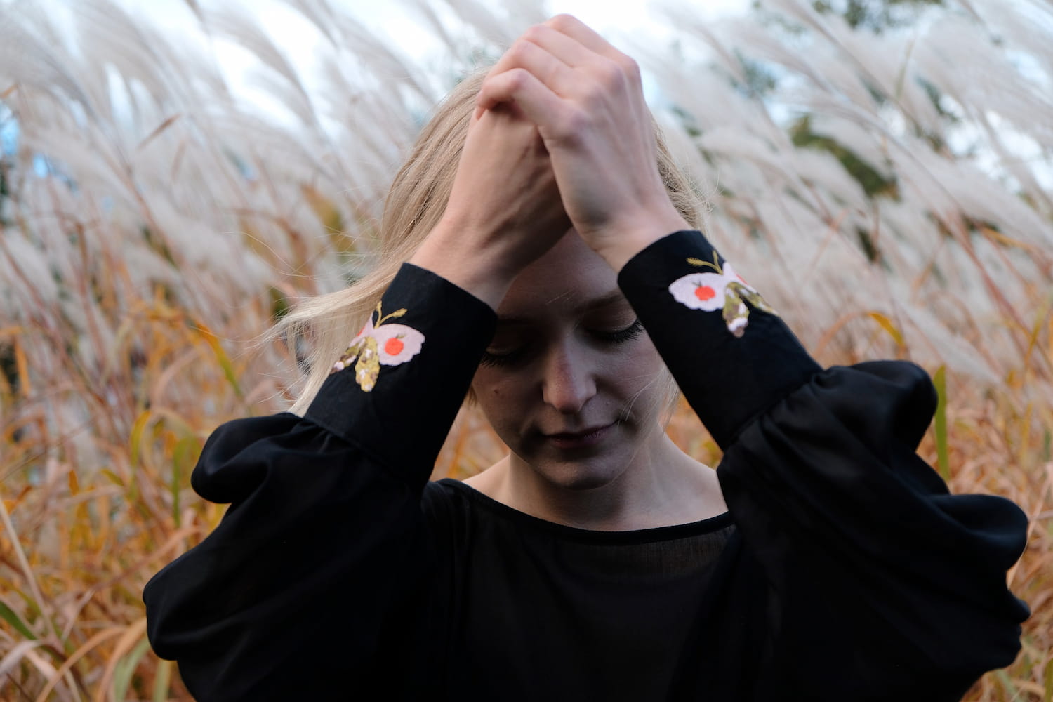 In an overcast field of tall grass, a person with blonde hair raises their hands above their head. They are wearing the Célia Butterfly Dress by Noémiah, which features exquisite floral embroidery on the sleeves and embroidered cuffs—the perfect complement to the soft, natural backdrop that accentuates its elegant details.