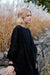 A person with short blond hair stands in front of a stone wall, wearing a long, loose Célia Butterfly Dress by Noémiah. The black silk cotton fabric features floral embroidery on the cuffs. Tall grass with fluffy white tops is in the background.