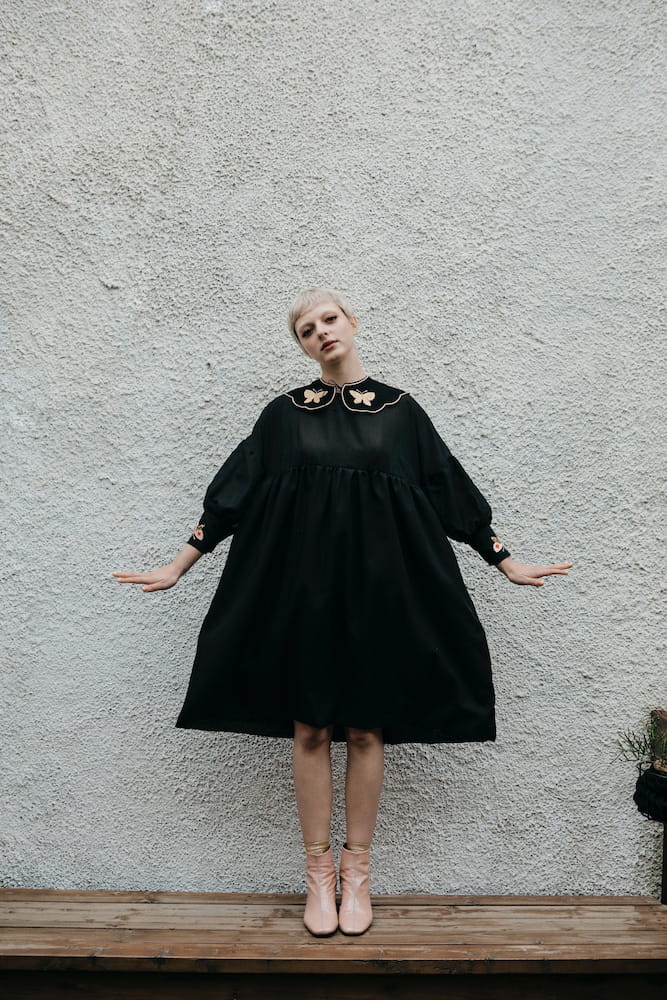 A person with short hair stands on a wooden platform against a textured wall, wearing a loose black Célia Butterfly dress by Noémiah, featuring delicate floral embroidery around the collar and cuffs. With their arms slightly outstretched, they wear light pink shoes that beautifully complement the silk cotton fabric of the outfit.