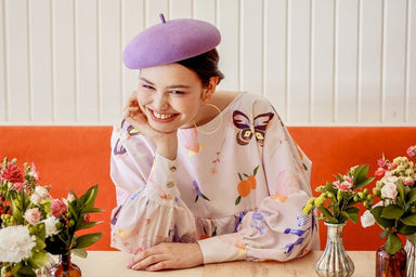 A person wearing the Célia Printed Dress from Noémiah and a purple beret, featuring the exclusive print from the Estée Preda collaboration, smiles while leaning on a table. The table is adorned with small vases containing colorful flowers. A bright orange booth and white wall are in the background.