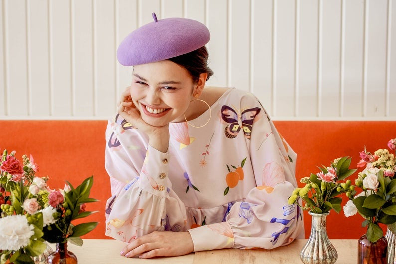 A person wearing the Célia Printed Dress from Noémiah and a purple beret, featuring the exclusive print from the Estée Preda collaboration, smiles while leaning on a table. The table is adorned with small vases containing colorful flowers. A bright orange booth and white wall are in the background.