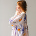 A woman with brown hair faces sideways, gently touching her face while wearing a vibrant Célia Printed Dress from Noémiah, featuring an exclusive print of floral and fruit patterns. This exquisite piece from the Estée Preda collaboration stands out beautifully against the plain light gray background.