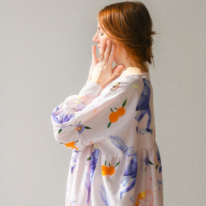 A woman with brown hair faces sideways, gently touching her face while wearing a vibrant Célia Printed Dress from Noémiah, featuring an exclusive print of floral and fruit patterns. This exquisite piece from the Estée Preda collaboration stands out beautifully against the plain light gray background.
