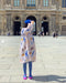 A person wearing a vibrant Célia Printed Dress by Noémiah, paired with a blue beret and pink shoes, stands on a cobblestone plaza in front of a grand, historic building adorned with arches and sculptures. They're facing away, possibly admiring the exclusive print from the Estée Preda collaboration.