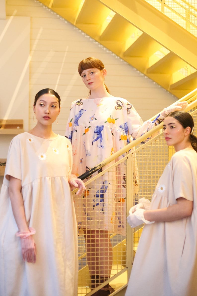 Three individuals dressed in chic, pastel-hued Célia Printed Dresses by Noémiah are positioned on a staircase. They exude confidence with two on the lower steps and one higher up, showcasing exclusive floral prints from the Estée Preda collaboration while holding onto the railing. The backdrop is softly illuminated with warm tones.