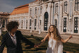 A couple joyfully holds hands and smiles while walking in front of an elegant, historic building with ornate architecture. The woman is wearing the flowing Céline Dress by Noémiah along with a veil, and the man is dressed smartly in a dark suit.