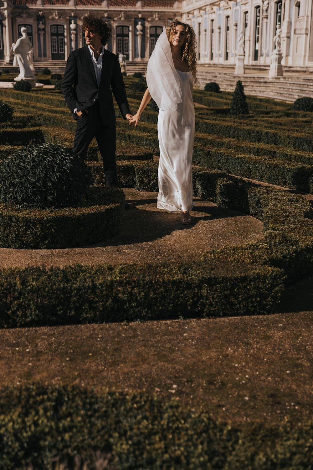 The bride, wearing the graceful Céline Dress by Noémiah, walks hand in hand with her partner through an ornate, manicured garden with hedges and topiaries. In the background stands a grand historical building adorned with statues and large windows.