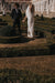 The bride, wearing the graceful Céline Dress by Noémiah, walks hand in hand with her partner through an ornate, manicured garden with hedges and topiaries. In the background stands a grand historical building adorned with statues and large windows.