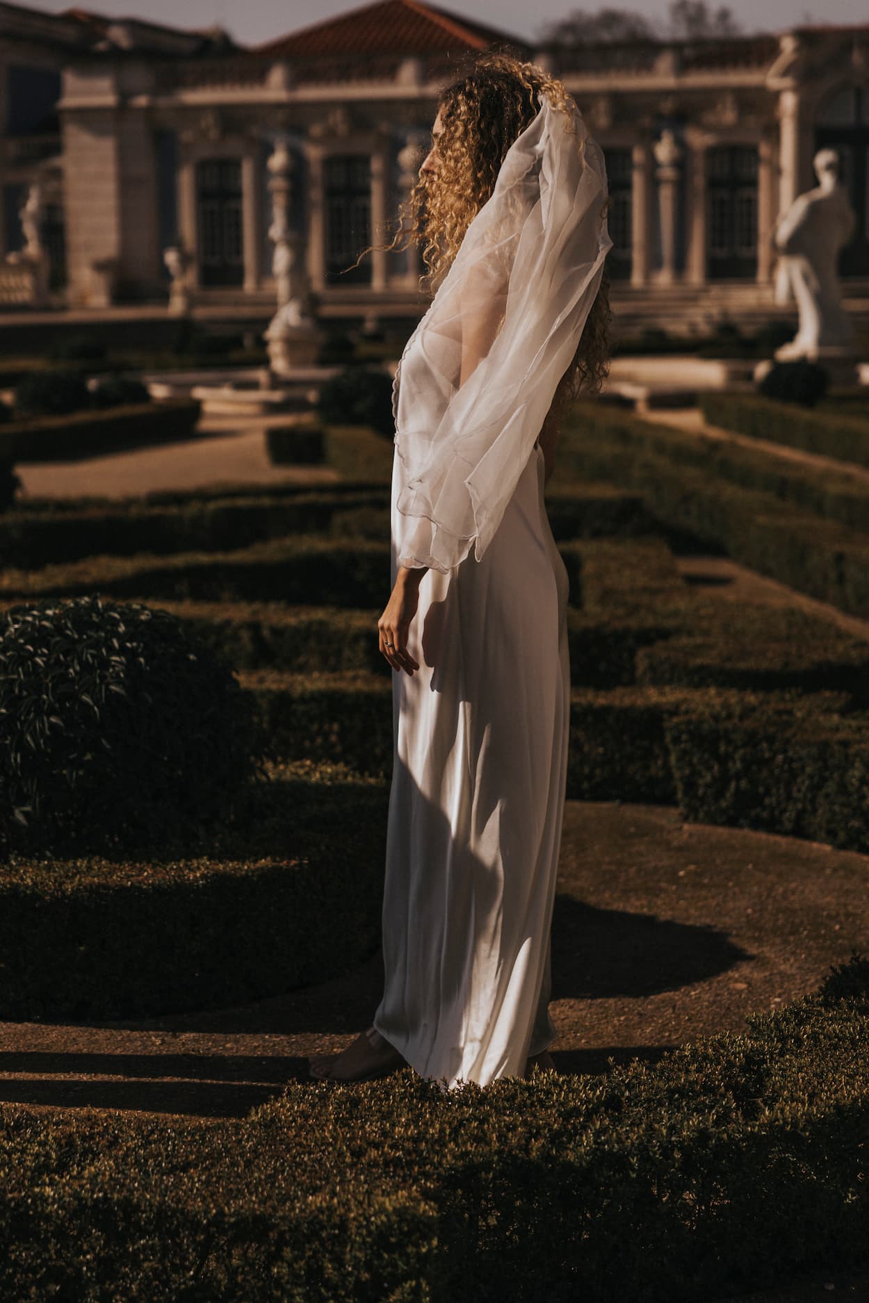 A woman with curly hair stands in a garden, wearing the elegant Céline Dress and a sheer shawl by Noémiah. Sculpted hedges and classical statues are visible in the background, all warmly lit by the sun.