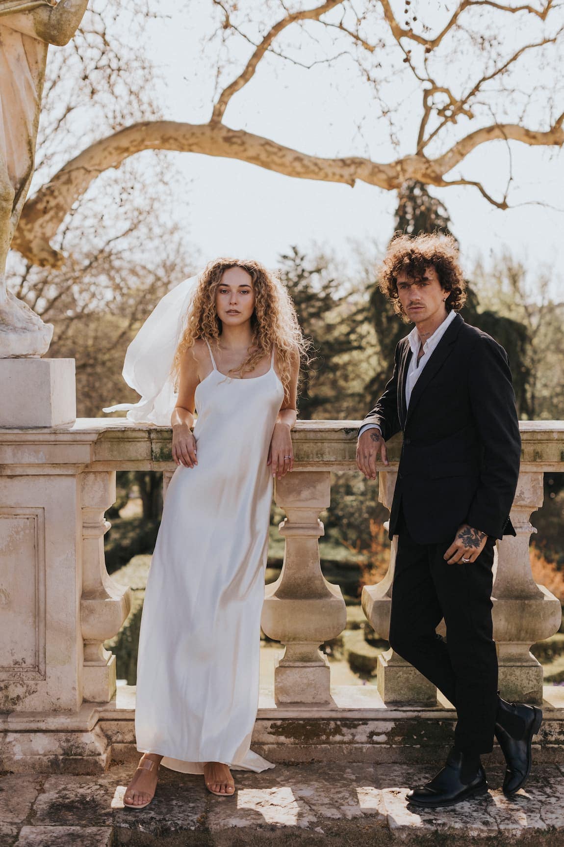 A woman in the elegant Céline Dress by Noémiah and a man in a black suit stand on a stone terrace. She leans against a balustrade while he stands beside her, both facing the camera, framed by bare trees in an outdoor garden setting.