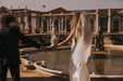 A bride in a stunning Noémiah Céline Dress walks hand in hand with the groom beside a large fountain in a garden. The white gown and veil beautifully complement his dark suit as they stand before a historic building adorned with statues.