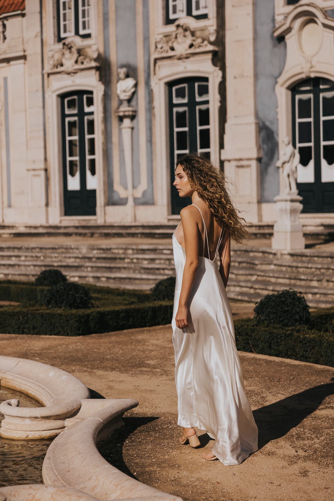 Wearing the enchanting Céline Dress from Noémiah, a woman gracefully strolled along a stone path in front of a historic building adorned with ornate windows and statues. The sunlight illuminated her curly hair and accentuated the gown’s luxurious texture, creating a captivating fusion of elegance and history.