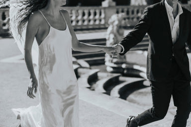 A black and white image of a couple holding hands and moving forward. The woman is wearing the elegant Noémiah Céline Dress, while the man is in a dark suit. They are near a fountain with stone sculptures in the background.