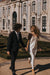 A couple joyfully walks hand in hand outside an ornate building with intricate architecture. The woman, barefoot in a flowing Céline Dress by Noémiah, adorned with a veil, is complemented by the man in a dark suit. They are surrounded by manicured gardens.