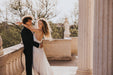 A couple embraces on a sunlit stone terrace. The woman wears the graceful Céline Dress by Noémiah, while the man is in a dark suit. Trees and classical columns create a serene backdrop.