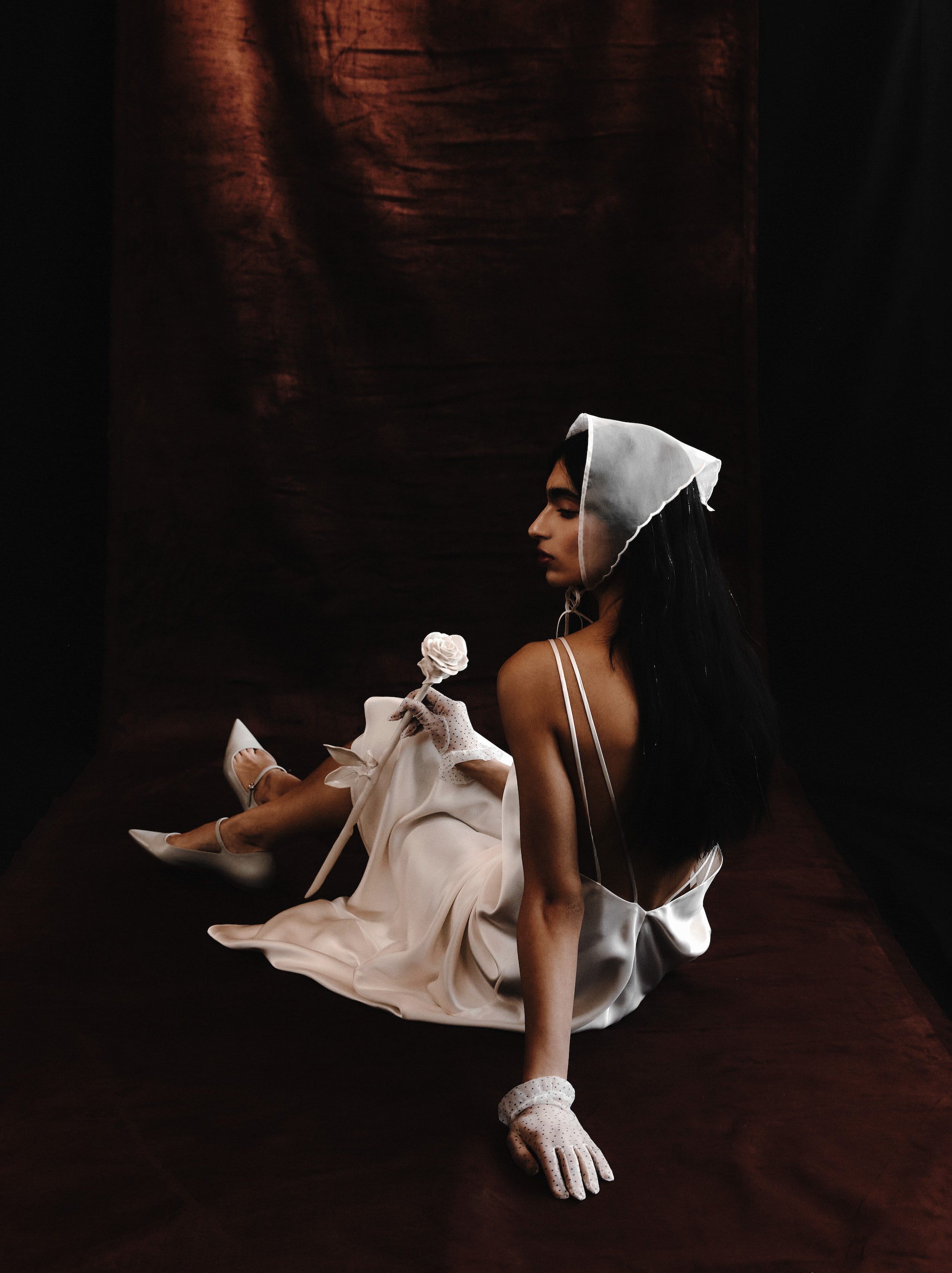 A person in the Noémiah Céline Dress, styled with sheer gloves and a white headscarf, sits before a dark brown backdrop holding a white rose, gazing contemplatively to the side.