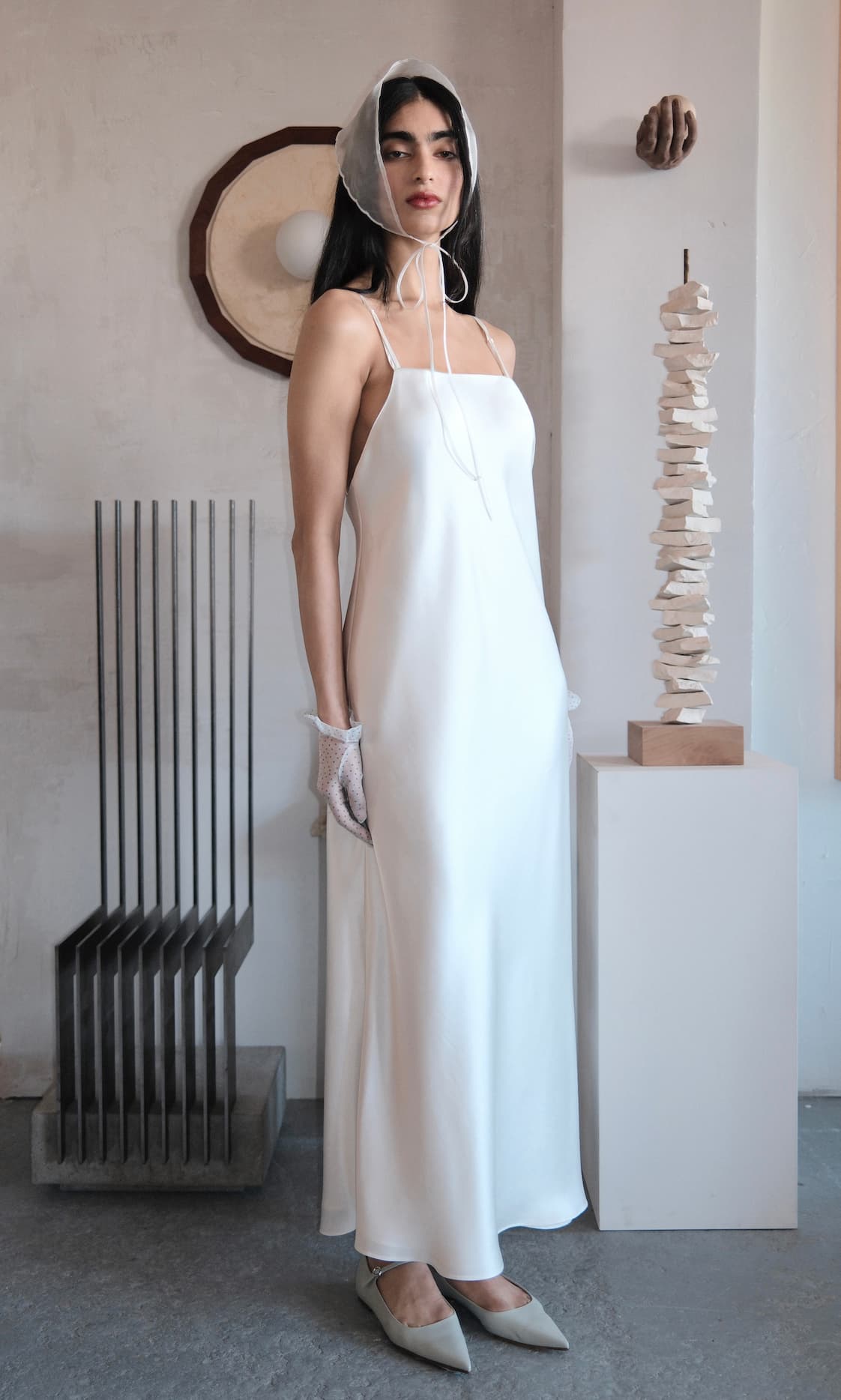 A woman in the Noémiah Céline Dress, styled with mesh gloves, pointed shoes, and a sheer bonnet, stands indoors by modern abstract sculptures against a neutral-toned wall.