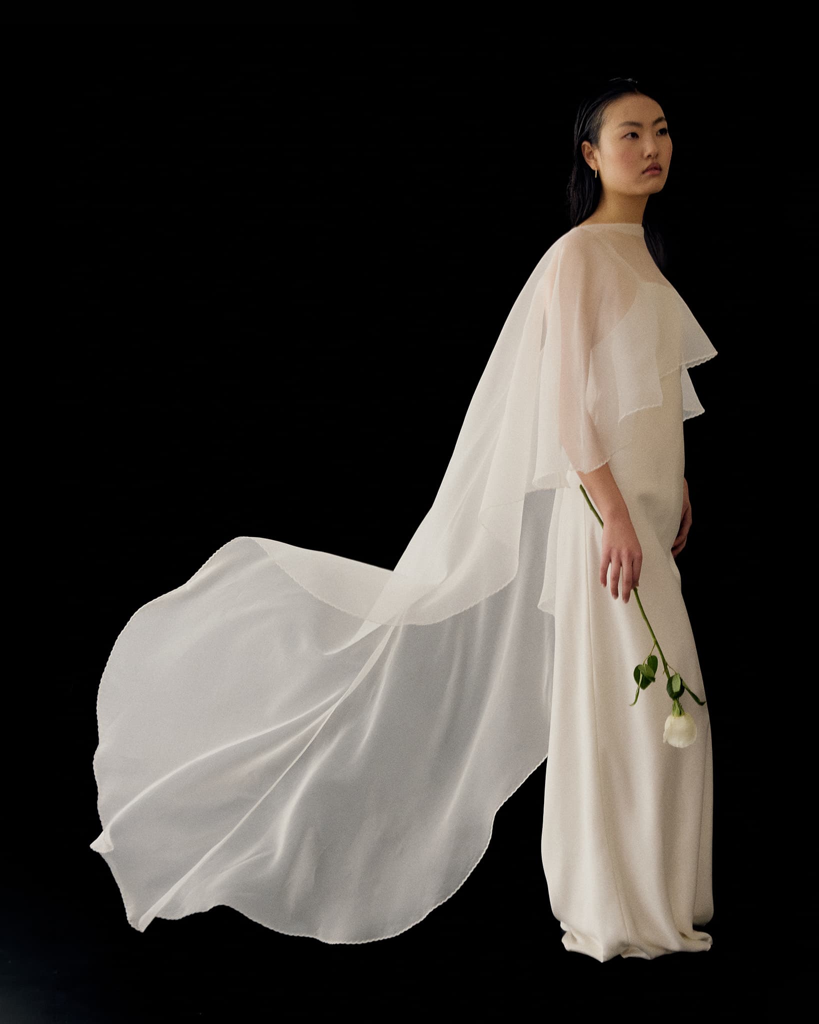 A woman in the Céline Dress by Noémiah stands against a black background, holding a white rose. The gown features long, layered, translucent fabric that trails gracefully behind her.