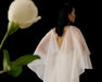 A woman with long dark hair wears the Noémiah Céline Dress—an elegant evening gown with a sheer white deep V-back—standing against a black backdrop, while a softly blurred white flower adds delicate contrast in the foreground.