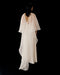 Against a black background, a person is shown from behind in the Noémiah Céline Dress—a long white evening gown with sheer, draped fabric flowing elegantly from the shoulders down the back.