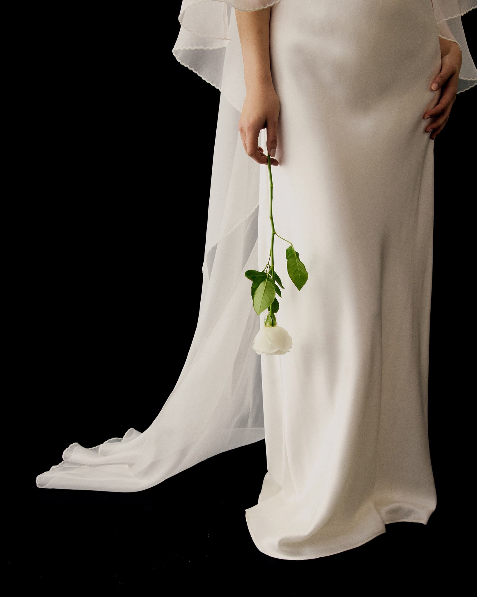 Wearing the Noémiah Céline Dress, a person holds a white rose by the stem, their lower half visible against a black background.