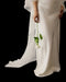 Wearing the Noémiah Céline Dress, a person holds a white rose by the stem, their lower half visible against a black background.
