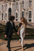 A couple joyfully walks hand in hand outside an ornate building with intricate architecture. The woman, barefoot in a flowing Céline Dress by Noémiah, adorned with a veil, is complemented by the man in a dark suit. They are surrounded by manicured gardens.