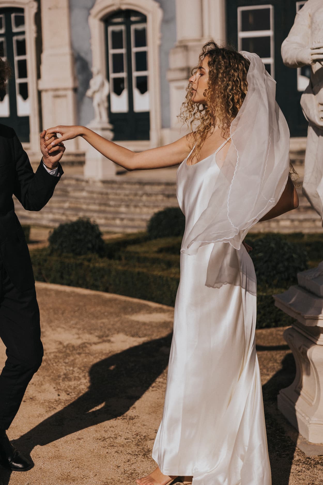 The bride, adorned in the elegant Céline Dress by Noémiah, holds hands with a groom, who is partially visible. They stand outside a building featuring large windows and ornate architecture.