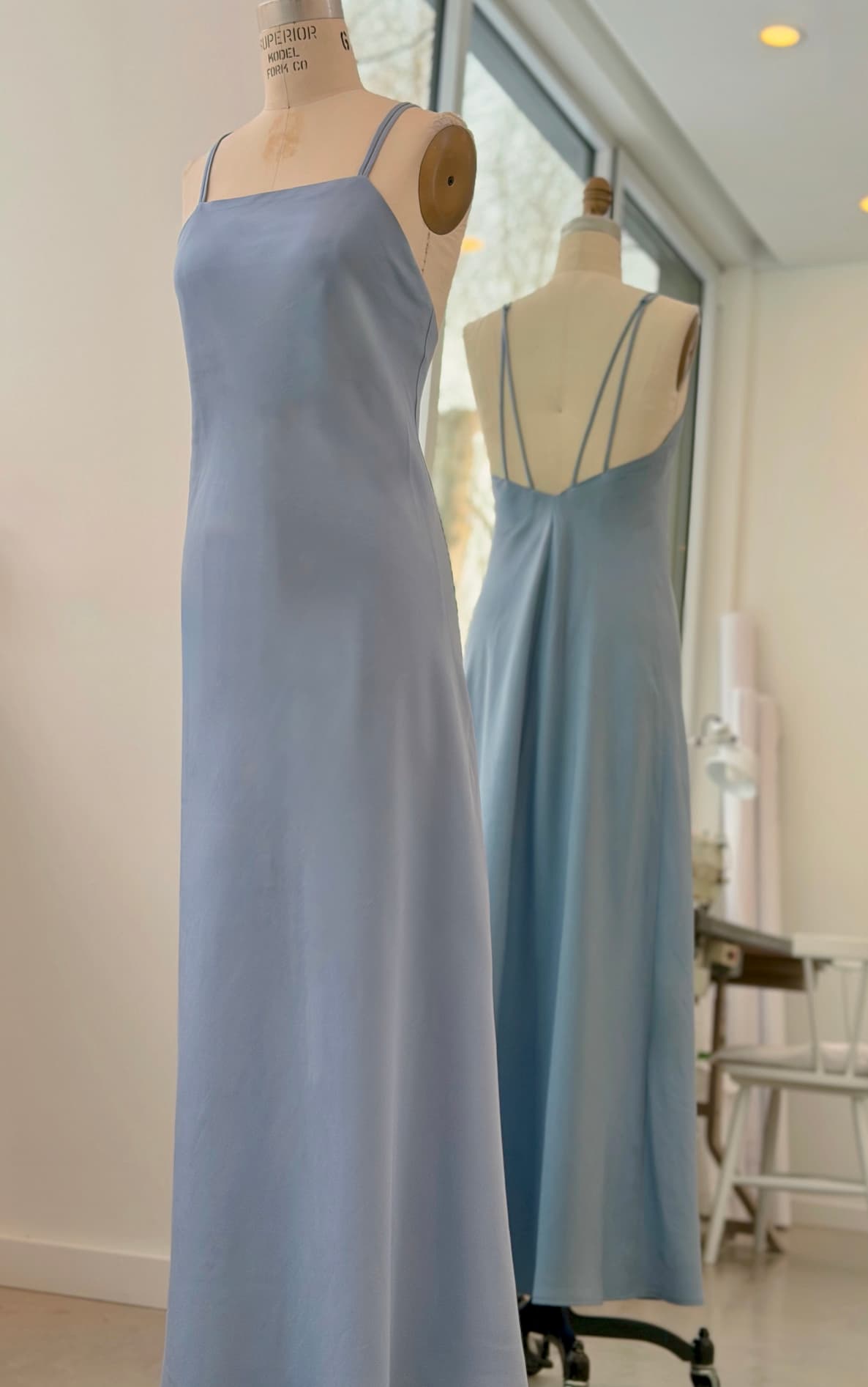 The Céline Lyocell Dress — Classic by Noémiah in light blue, featuring thin straps and a strappy back, is displayed on a dress form before a mirror in a bright, modern studio.