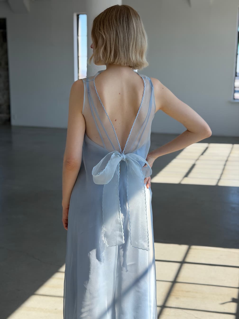 A woman with short blond hair stands indoors, wearing the Noémiah Céline Lyocell Dress — Classic. The light blue sustainable dress features a low, sheer back and a large bow at the waist as sunlight streams onto the concrete floor.
