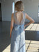 A woman with short blond hair stands indoors, wearing the Noémiah Céline Lyocell Dress — Classic. The light blue sustainable dress features a low, sheer back and a large bow at the waist as sunlight streams onto the concrete floor.
