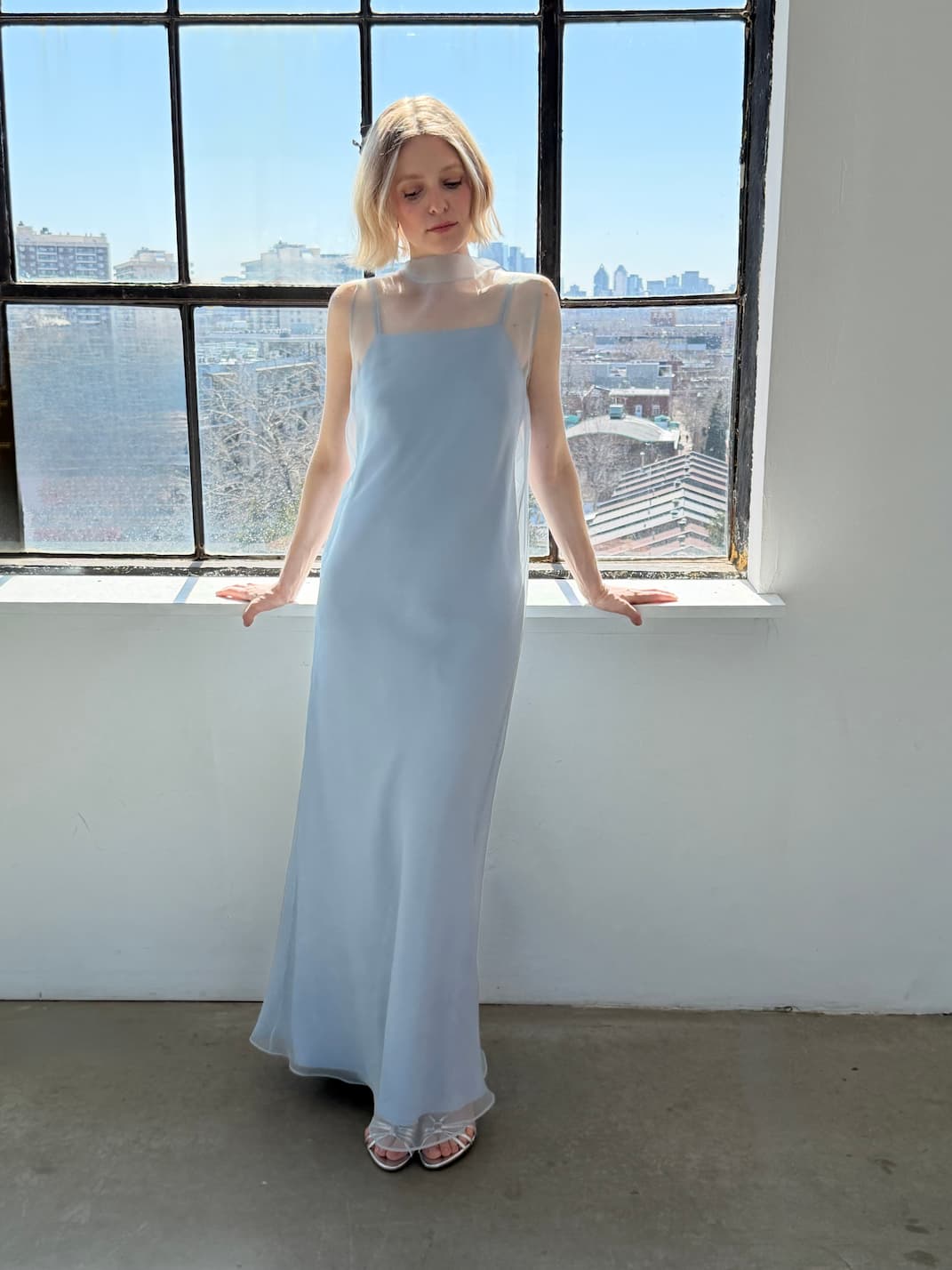A woman in the Noémiah Céline Lyocell Dress — Classic stands by a large window, sunlight streaming in as she looks down, hands resting on the sill. City buildings and blue sky are visible outside.