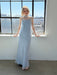 A woman in the Noémiah Céline Lyocell Dress — Classic stands by a large window, sunlight streaming in as she looks down, hands resting on the sill. City buildings and blue sky are visible outside.