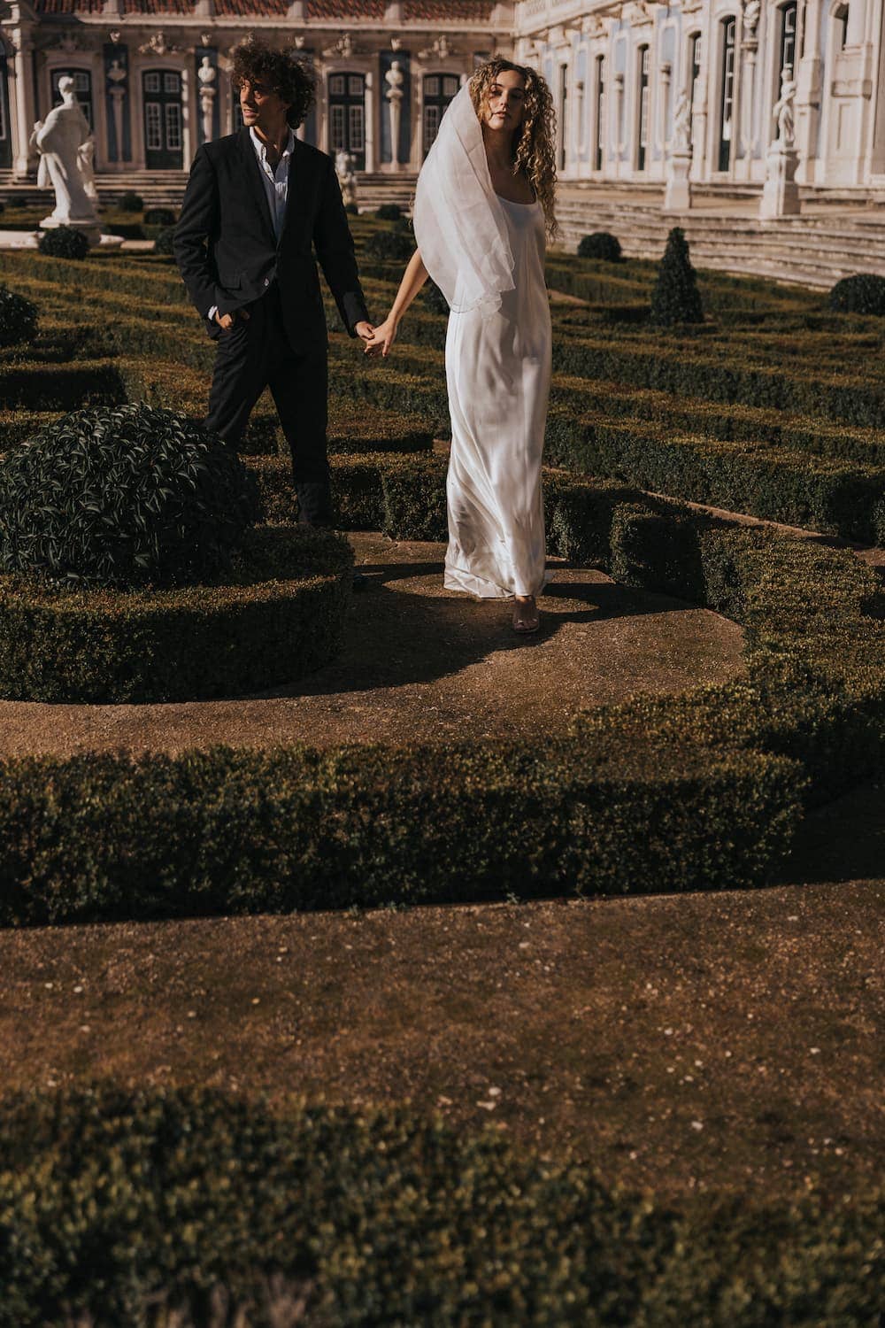 A bride in the Noémiah Céline Silk Satin Dress — Bridal Edit and veil holds hands with a groom in a black suit as they walk through a manicured garden maze before an elegant, historic building.