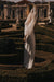 A woman in the Noémiah Céline Silk Satin Dress — Bridal Edit stands in profile amid sculpted hedges and classical statues in a sunlit formal garden, her gown’s sheer sleeves and lustrous fabric highlighted by strong shadows.