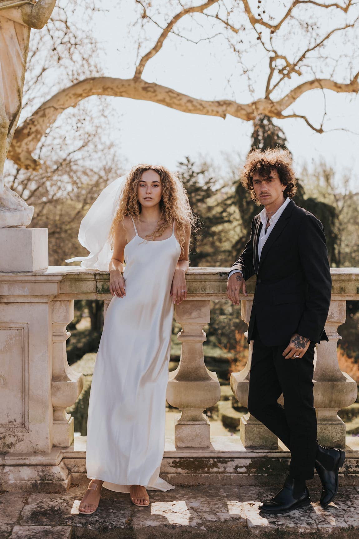 Wearing the Noémiah Céline Silk Satin Dress — Bridal Edit and veil, the bride stands beside a groom in a black suit on a sunny stone balcony, surrounded by trees for a romantic outdoor setting.