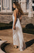 A woman with long curly hair wears the Céline Silk Satin Dress — Bridal Edit by Noémiah with beige heels, standing outdoors in front of stone steps and an elegant building.