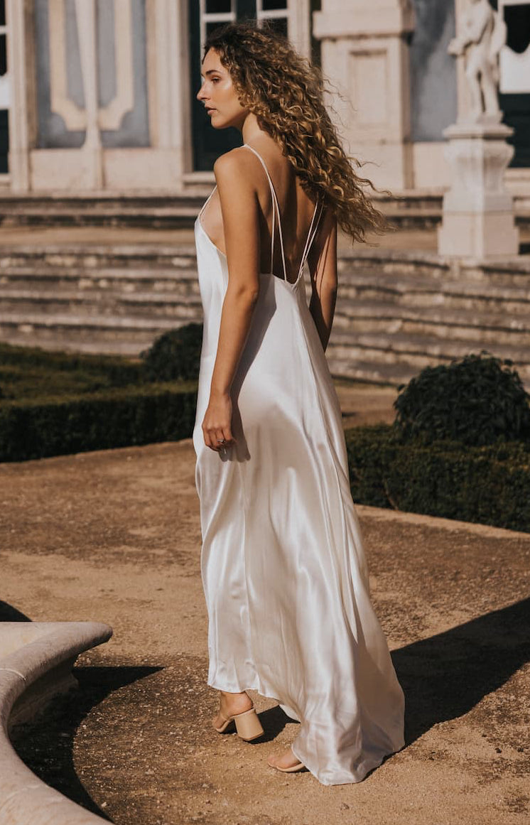 A woman with long curly hair wears the Céline Silk Satin Dress — Bridal Edit by Noémiah with beige heels, standing outdoors in front of stone steps and an elegant building.