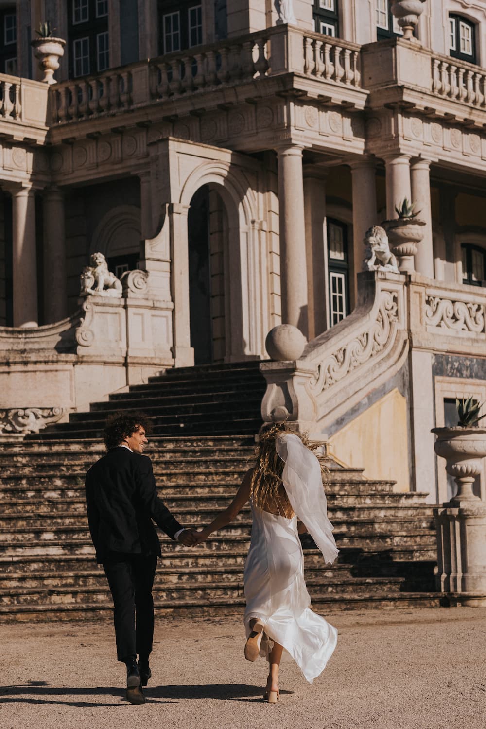 A couple, in wedding attire, ascend grand stone steps. The bride wears the Céline Silk Satin Dress by Noémiah and a veil; the groom is in a black suit, surrounded by ornate architecture and statues.
