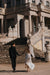 A couple, in wedding attire, ascend grand stone steps. The bride wears the Céline Silk Satin Dress by Noémiah and a veil; the groom is in a black suit, surrounded by ornate architecture and statues.