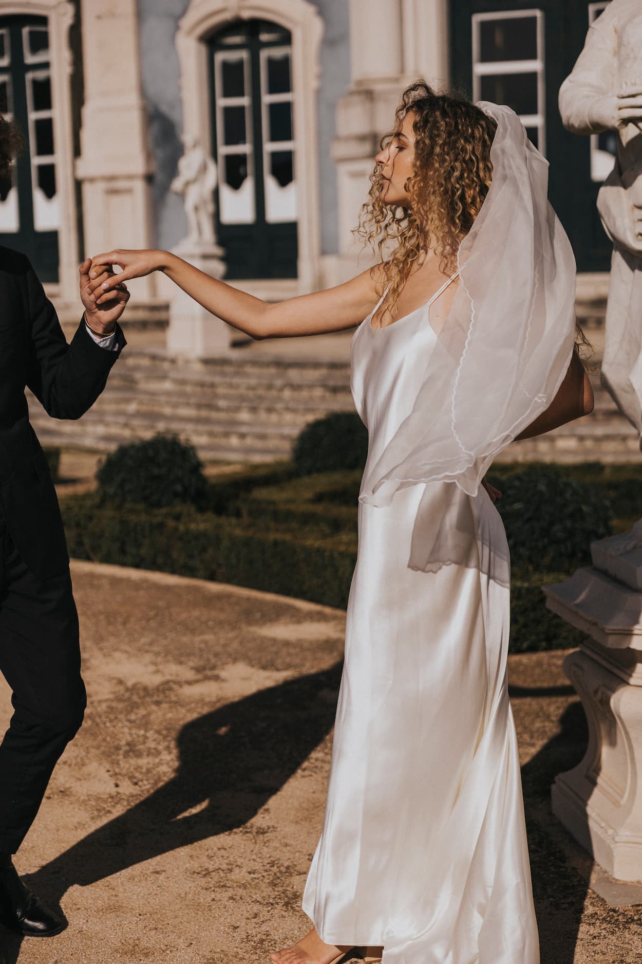 A bride in a Noémiah Céline Silk Satin Dress and veil holds hands with someone in a black suit outdoors, standing before a grand building with large windows and statues.