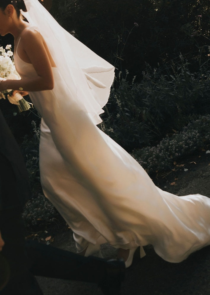 A bride in a Céline Silk Satin Dress — Bridal Edit by Noémiah walks outdoors, holding a white bouquet, sunlight highlighting the luxurious silk satin gown and her flowing veil.
