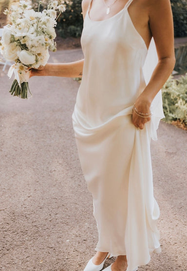 A woman in the Noémiah Céline Silk Satin Dress — Bridal Edit holds white flowers in one hand and gathers her elegant midi dress with the other while standing on a sunlit path.