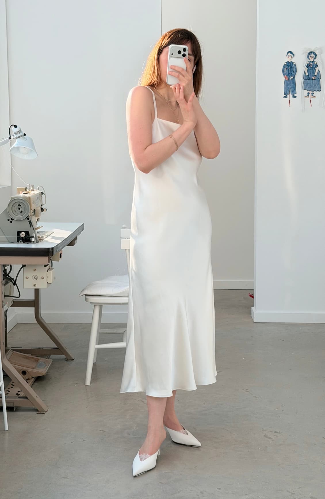 A woman wearing the Noémiah Céline Silk Satin Dress — Bridal Edit and white pointed shoes takes a mirror selfie in a bright room with a sewing machine, chair, and fashion sketches on the wall.