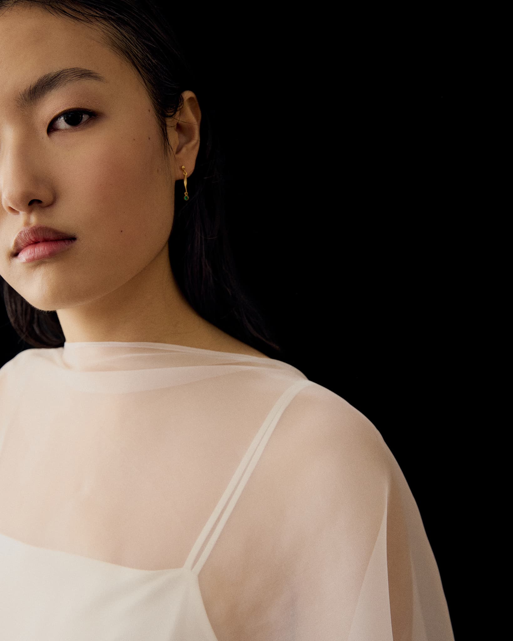 A woman with long dark hair wears the Noémiah Céline Silk Satin Dress — Bridal Edit and a gold earring, posed against a black background with only the left side of her face and upper body visible.