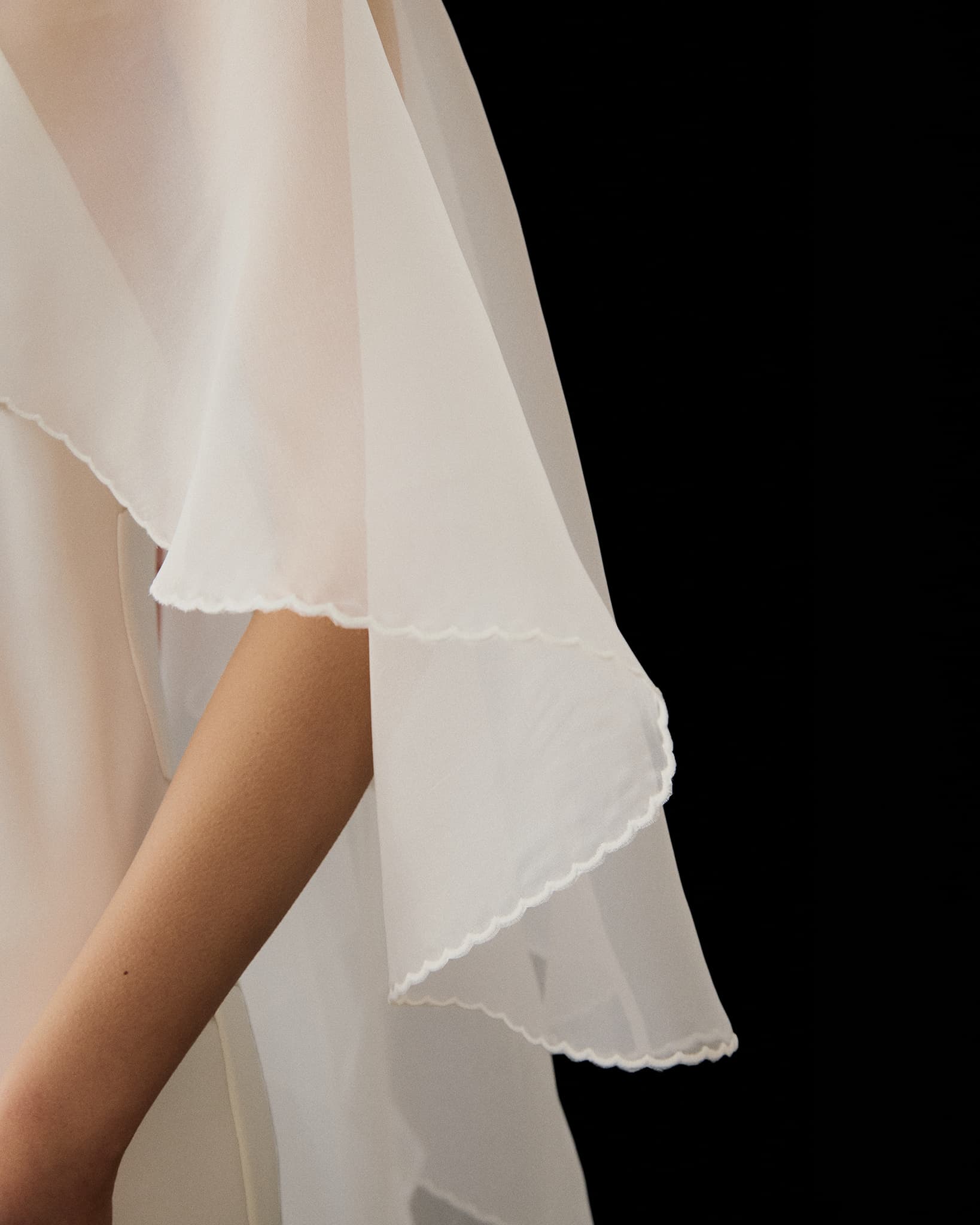 A close-up of a person’s arm in the Noémiah Céline Dress—sheer, flowing white fabric with scalloped edges drapes elegantly against a black background, creating soft, delicate folds reminiscent of an evening gown.