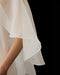 A close-up of a person’s arm in the Noémiah Céline Dress—sheer, flowing white fabric with scalloped edges drapes elegantly against a black background, creating soft, delicate folds reminiscent of an evening gown.